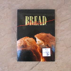 May include: A cookbook titled "Bread Electric" with a black cover and gold lettering. The cover features a photo of two halves of a loaf of bread. A red line runs across the title. A small sticker is on the bottom right corner.