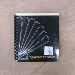 May include: A spiral-bound cookbook titled "Tidewater on the Half Shell" with a black cover featuring a white fan design. The book has a white spine and is on a light brown surface.