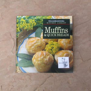 May include: A cookbook titled "Muffins & Quick Breads" from the Williams-Sonoma Kitchen Library. The cover features a photo of golden muffins on a plate with green leaves and yellow flowers. The book is on a textured, brown surface.