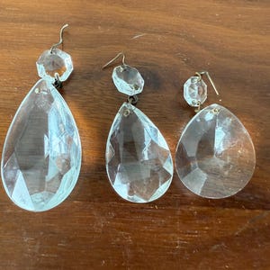 May include: Three clear, faceted crystal chandelier pendants with metal hooks. The pendants are teardrop-shaped, with a smaller, octagonal crystal above each. The crystals are displayed on a wooden surface.