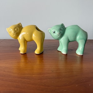 May include: Two ceramic cat figurines, one yellow and one green, standing on a wooden surface. Both cats have black eyes and a simple design.