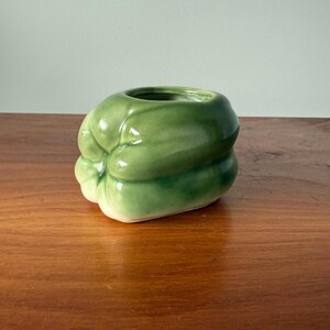 May include: A ceramic, green bell pepper-shaped container. The glossy, light green exterior has darker green shading, with a white base. The container has a circular opening at the top. The object is on a wooden surface.