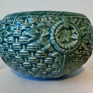 Puede incluir: Un cuenco de cerámica verde azulado con forma de cesta y textura tejida. El cuenco presenta un diseño detallado con detalles florales y circulares en relieve. Es una pieza decorativa, adecuada para diversos usos.