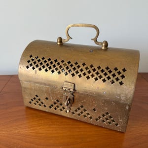Puede incluir: Una caja de latón con tapa abatible y una cerradura. La caja tiene un diseño decorativo perforado y un asa en la parte superior.