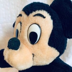 May include: Close-up of a plush Mickey Mouse toy with black fur, large white eyes, and a black nose. The toy is looking to the right.