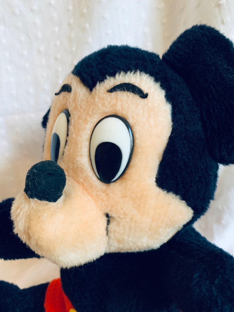 May include: Close-up of a plush Mickey Mouse toy with black fur, large white eyes, and a black nose. The toy is looking to the right.