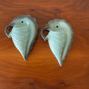 May include: Two teal green ceramic wall planters shaped like leaves. The planters have a textured surface and a hole at the top for drainage.