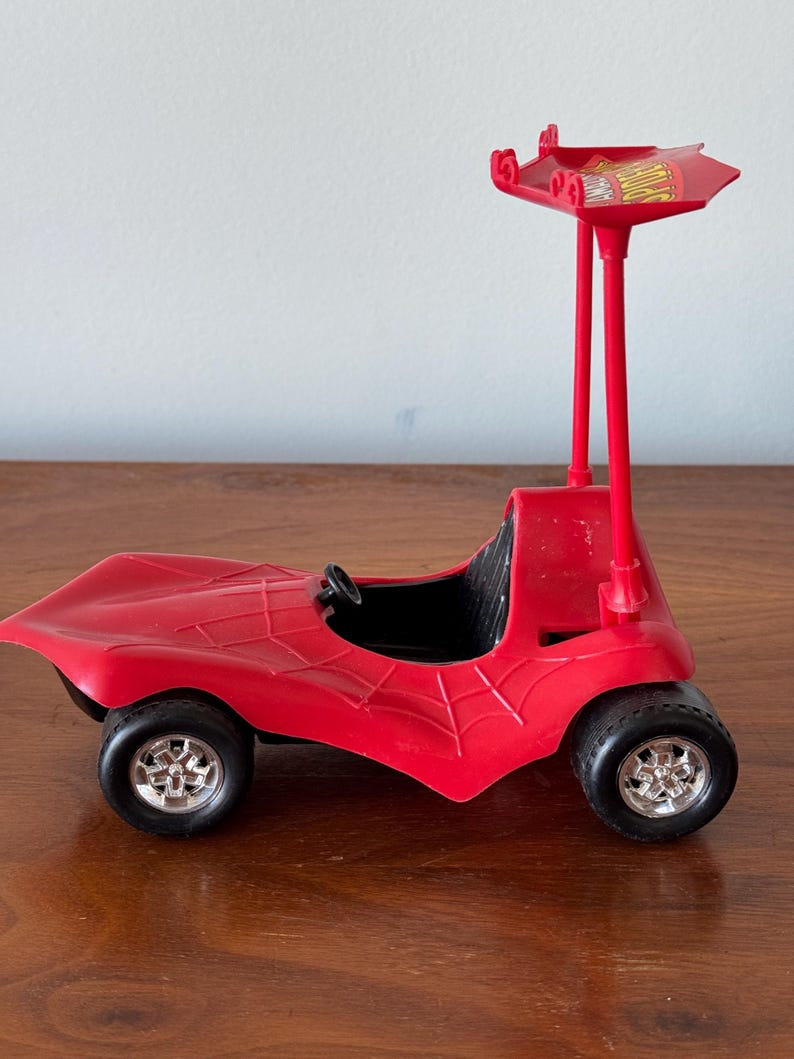 Pu&ograve; includere: Macchina giocattolo di plastica rossa con un design a ragnatela e un'antenna rossa alta con un cartello giallo che dice "Spider-Man".