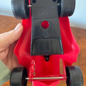 Pu&ograve; includere: Macchina da corsa giocattolo rossa con ruote nere e un asse argentato. La macchina ha una base in plastica nera con il testo "&copy; 1976  MIKE  JOYNER  ALL RIGHTS RESERVED  NEW YORK, N.Y. 10022  MADE IN TAIWAN" stampato sul fondo.