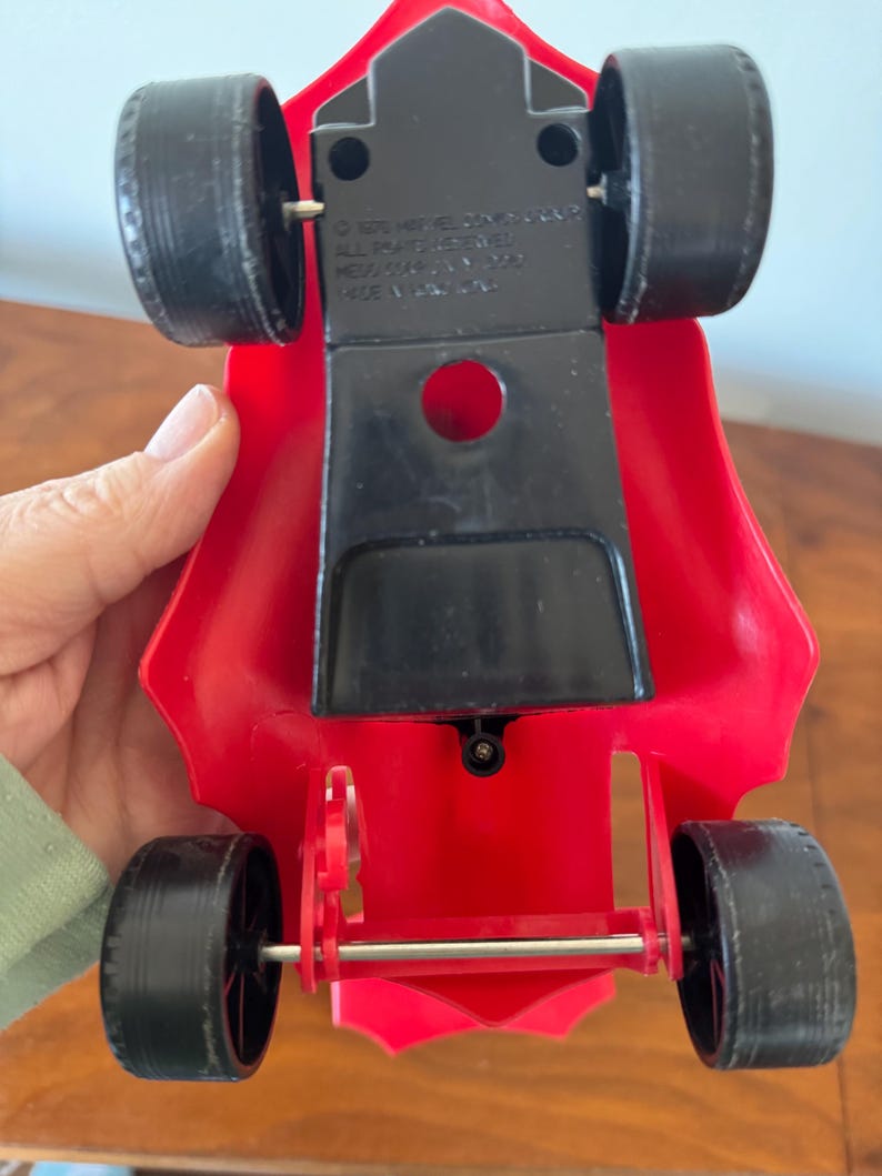 Pu&ograve; includere: Macchina da corsa giocattolo rossa con ruote nere e un asse argentato. La macchina ha una base in plastica nera con il testo "&copy; 1976  MIKE  JOYNER  ALL RIGHTS RESERVED  NEW YORK, N.Y. 10022  MADE IN TAIWAN" stampato sul fondo.