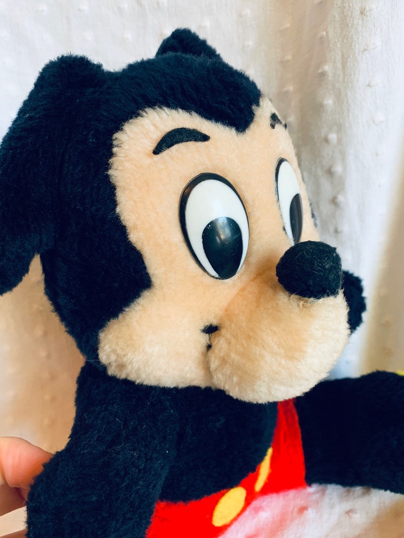 May include: A vintage plush Mickey Mouse toy with black fur, large black eyes, a brown nose, and red shorts with yellow buttons.