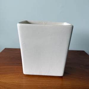 May include: A square, white ceramic planter with a slightly tapered design. The planter is empty and sits on a wooden surface. The background is a solid light blue.