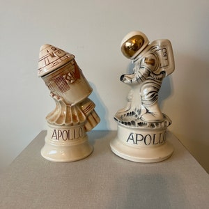 May include: Two ceramic figurines: a rocket and an astronaut. The rocket is beige and brown with the word "APOLLO" at the base. The astronaut is white with a gold helmet and the word "APOLLO" at the base. The figurines are on a gray surface.