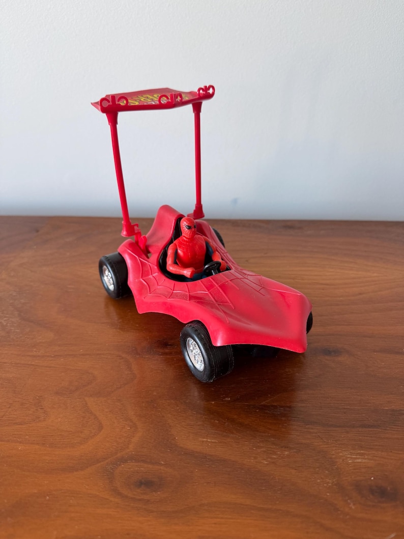 Pu&ograve; includere: Macchina giocattolo rossa con una figura di Spider-Man al volante. La macchina ha una cappotta rossa e ruote nere.