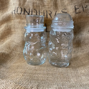 May include: Two clear glass jars shaped like snowmen. The jars have a textured surface and are decorated with raised details. The jars are sitting on a burlap background.