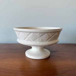 Puede incluir: Un cuenco con pedestal de color crema con un patrón de diamantes alrededor del borde. El cuenco tiene una base ancha y poco profunda y un tallo corto y redondeado. El cuenco está hecho de cerámica o porcelana y se encuentra sobre una superficie de madera.