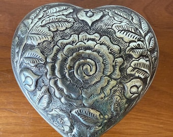 Puede incluir: Caja plateada en forma de corazón con intrincados diseños florales y de hojas. La caja presenta un diseño central de rosa rodeado de follaje detallado. El metal tiene una apariencia ligeramente envejecida, lo que aumenta su encanto vintage.