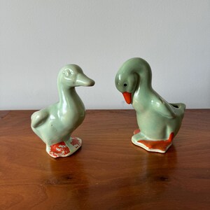 May include: Two ceramic duck figurines, both in a light green glaze with orange accents. The ducks are facing different directions, one with its head down and the other with its head up.