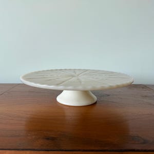 Puede incluir: Un soporte para pasteles de cerámica color crema, con un borde festoneado y un patrón decorativo de estrella en la parte superior. El soporte tiene una base corta y redondeada y se asienta sobre una superficie de madera oscura.