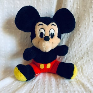 May include: A plush black Mickey Mouse toy with large, round eyes and a red and yellow outfit. The toy is sitting on a white textured surface.