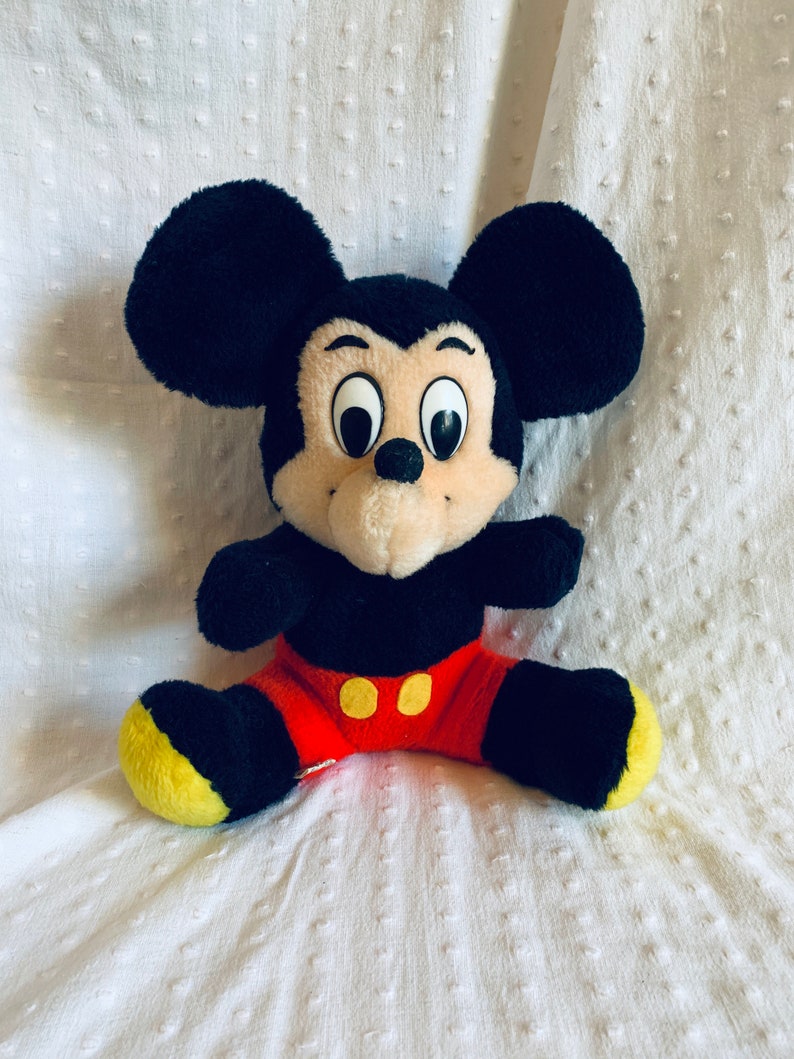 May include: A plush black Mickey Mouse toy with large, round eyes and a red and yellow outfit. The toy is sitting on a white textured surface.