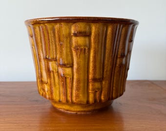 May include: A ceramic planter with a textured bamboo design in shades of brown and yellow. The planter has a wide opening and a slightly tapered shape. The rim is a darker brown, and the planter sits on a wooden surface.