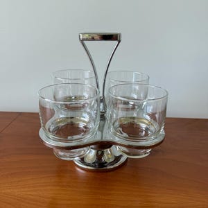 Könnte beinhalten: Ein silberfarbenes Serviertablett mit vier transparenten Trinkgläsern. Das Tablett hat einen Griff und einen kreisförmigen Sockel. Die Gläser sind kreisförmig angeordnet, ideal zum Servieren von Getränken oder kleinen Portionen.