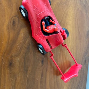 Pu&ograve; includere: Un'auto giocattolo rossa con un design a ragnatela e una action figure rossa e blu di Spider-Man sul sedile del conducente.