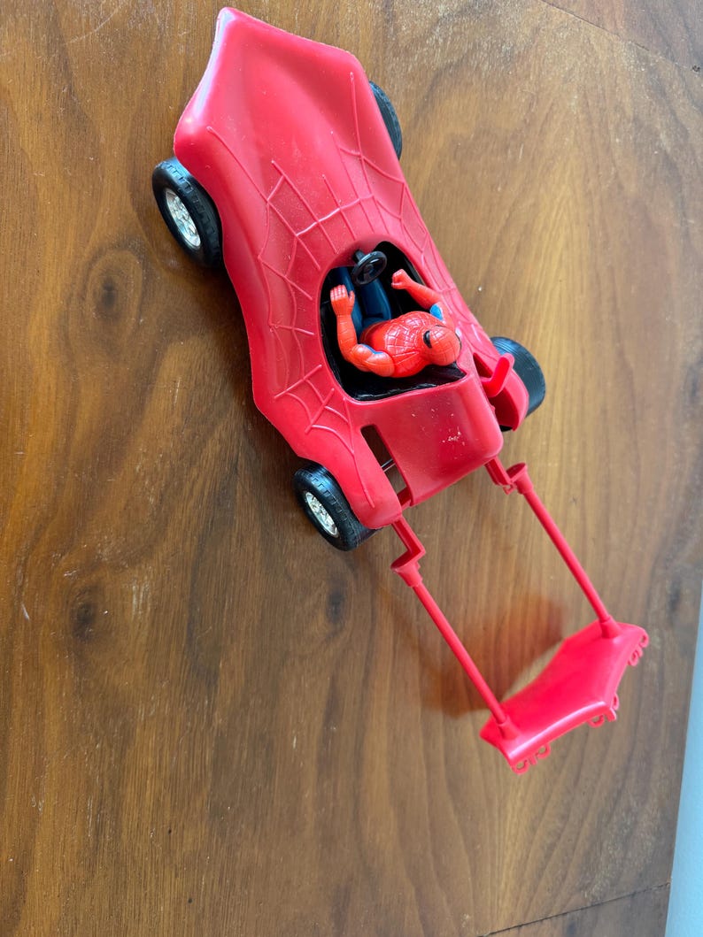 Pu&ograve; includere: Un'auto giocattolo rossa con un design a ragnatela e una action figure rossa e blu di Spider-Man sul sedile del conducente.