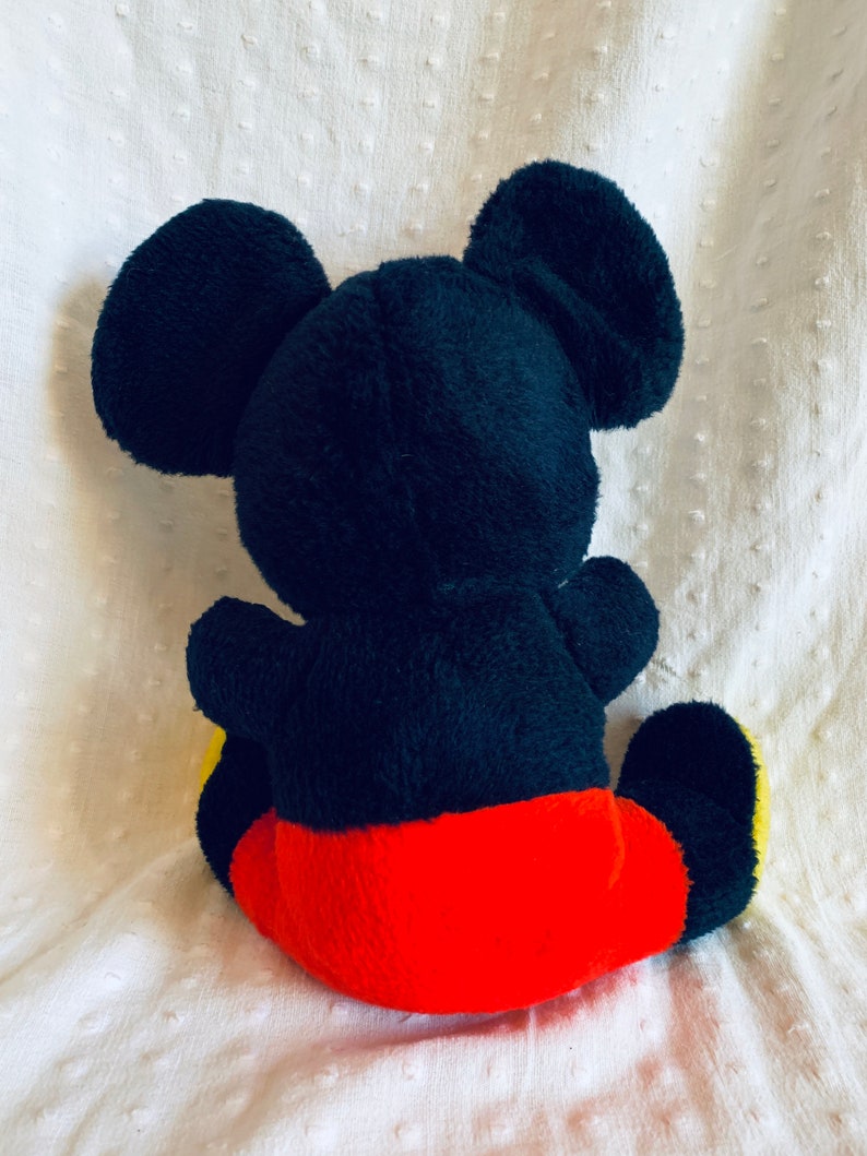 May include: A black plush Mickey Mouse toy with red shorts and yellow shoes. The toy is sitting on a white surface.