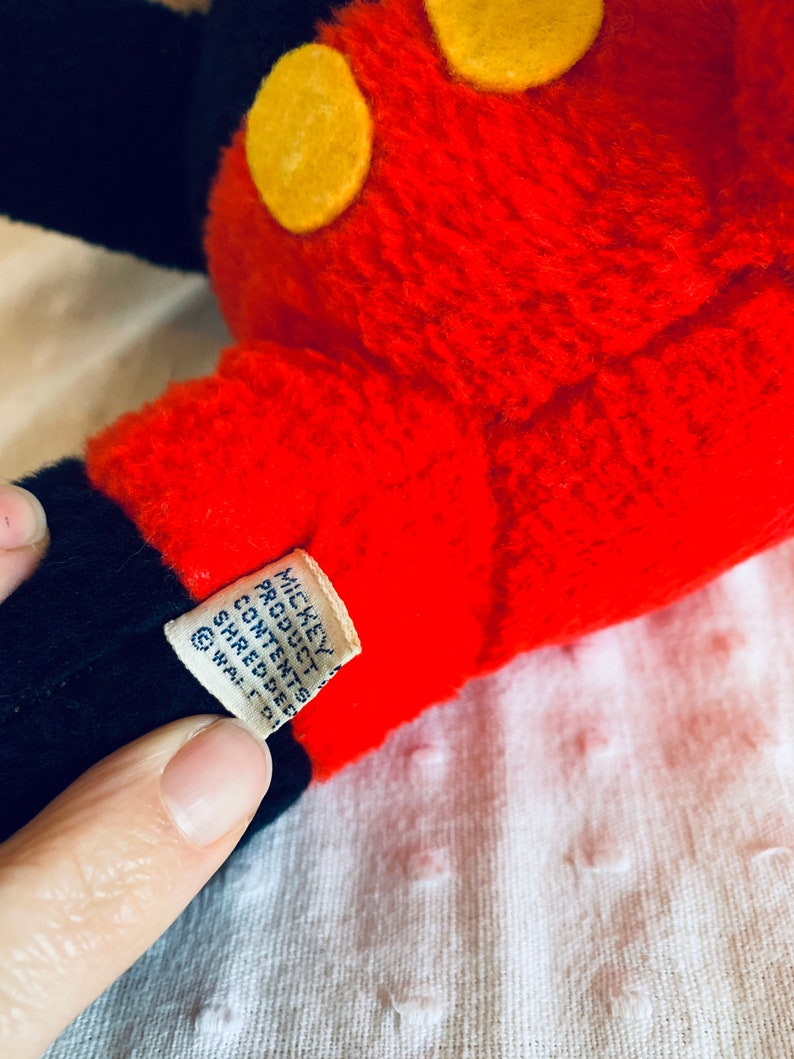May include: Close-up of a red and black Mickey Mouse plush toy with yellow felt circles. The tag reads "Mickey Product Contents Shredded  &copy; Walt D.  ",