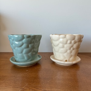 May include: Two ceramic flower pots with matching saucers. The pot on the left is a light blue color and the pot on the right is white. Both pots have a textured, raised design.