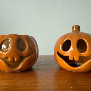 May include: Two glossy orange ceramic pumpkin-shaped containers with carved faces featuring wide smiles, two teeth, and hollow eyes. The pumpkin on the left is open at the top, while the pumpkin on the right has a stem and a hollow interior.