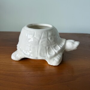 May include: A white ceramic turtle-shaped planter. The turtle has a textured shell, four small feet, and a head extending forward. The planter has a circular opening on top for plants or small items. The turtle is sitting on a wooden surface.