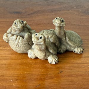 May include: A set of three small, light brown stone-like turtle figurines. One turtle is emerging from a cracked eggshell, while the other two are positioned in front, featuring textured shells, small black eyes, and detailed claws on their feet.