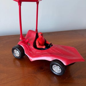 Pu&ograve; includere: Un'auto giocattolo rossa con un design a ragnatela nera e una tettoia rossa. Una action figure rossa e blu &egrave; seduta sul sedile del conducente.