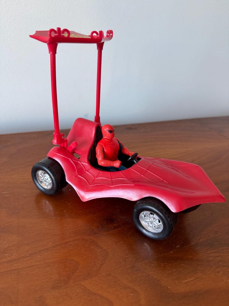 Pu&ograve; includere: Un'auto giocattolo rossa con un design a ragnatela nera e una tettoia rossa. Una action figure rossa e blu &egrave; seduta sul sedile del conducente.
