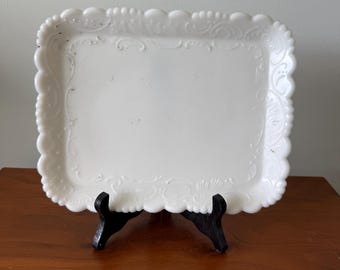 May include: A white, square serving tray with a scalloped edge and ornate, embossed floral designs. The tray is displayed on a black stand, showcasing its decorative details. A vintage piece for serving or display.
