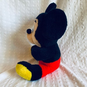 May include: A black and red plush Mickey Mouse toy sitting on a white textured surface. The toy is facing to the right and has a yellow and black shoe.