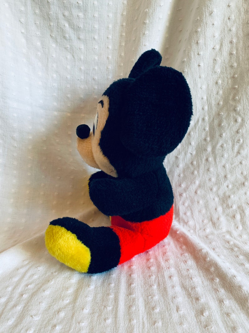 May include: A black and red plush Mickey Mouse toy sitting on a white textured surface. The toy is facing to the right and has a yellow and black shoe.