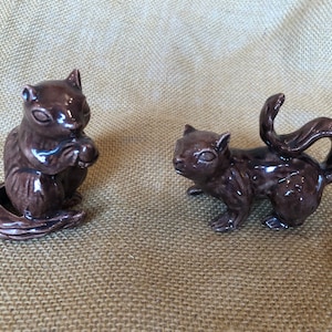 May include: Two brown ceramic squirrel figurines. One squirrel is sitting and holding a nut, while the other is standing with its tail curved upwards. The figurines are detailed and have a glossy finish. They are likely decorative items.