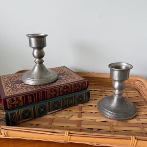 May include: Two antique pewter candlesticks with a classic design. The candlesticks are displayed on a woven tray with two vintage books. The books have decorative covers in burgundy and teal.