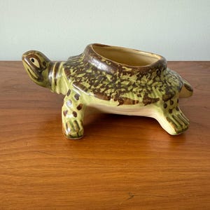 May include: A green ceramic turtle planter with brown spots. The turtle is in a walking position with its head turned to the side. The planter is empty.