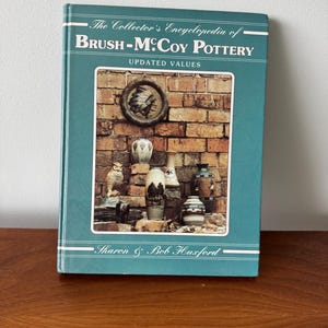 May include: The Collector's Encyclopedia of Brush-McCoy Pottery book with updated values by Sharon & Bob Huxford. The cover features a brick wall with a Native American headdress and several pottery pieces.