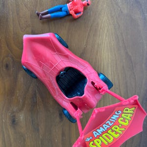 Pu&ograve; includere: Un'auto giocattolo rossa con un design a ragnatela e un cartello che dice "The Amazing Spider-Car". Una action figure rossa e blu di Spider-Man &egrave; sdraiata su una superficie di legno.