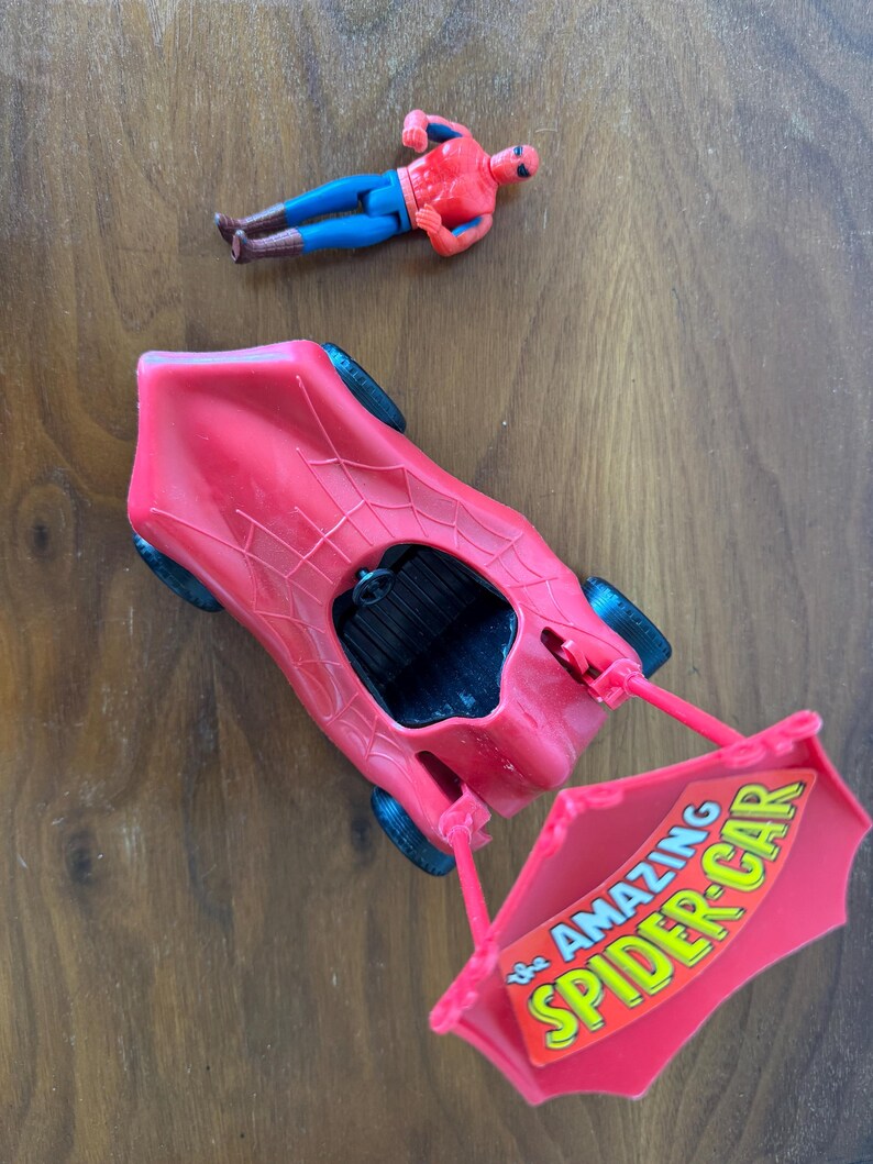Pu&ograve; includere: Un'auto giocattolo rossa con un design a ragnatela e un cartello che dice "The Amazing Spider-Car". Una action figure rossa e blu di Spider-Man &egrave; sdraiata su una superficie di legno.
