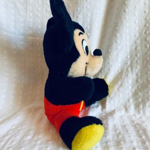 May include: A plush Mickey Mouse toy sitting on a white surface. The toy is black with red shorts and yellow shoes. Mickey Mouse has a black nose and large black ears.