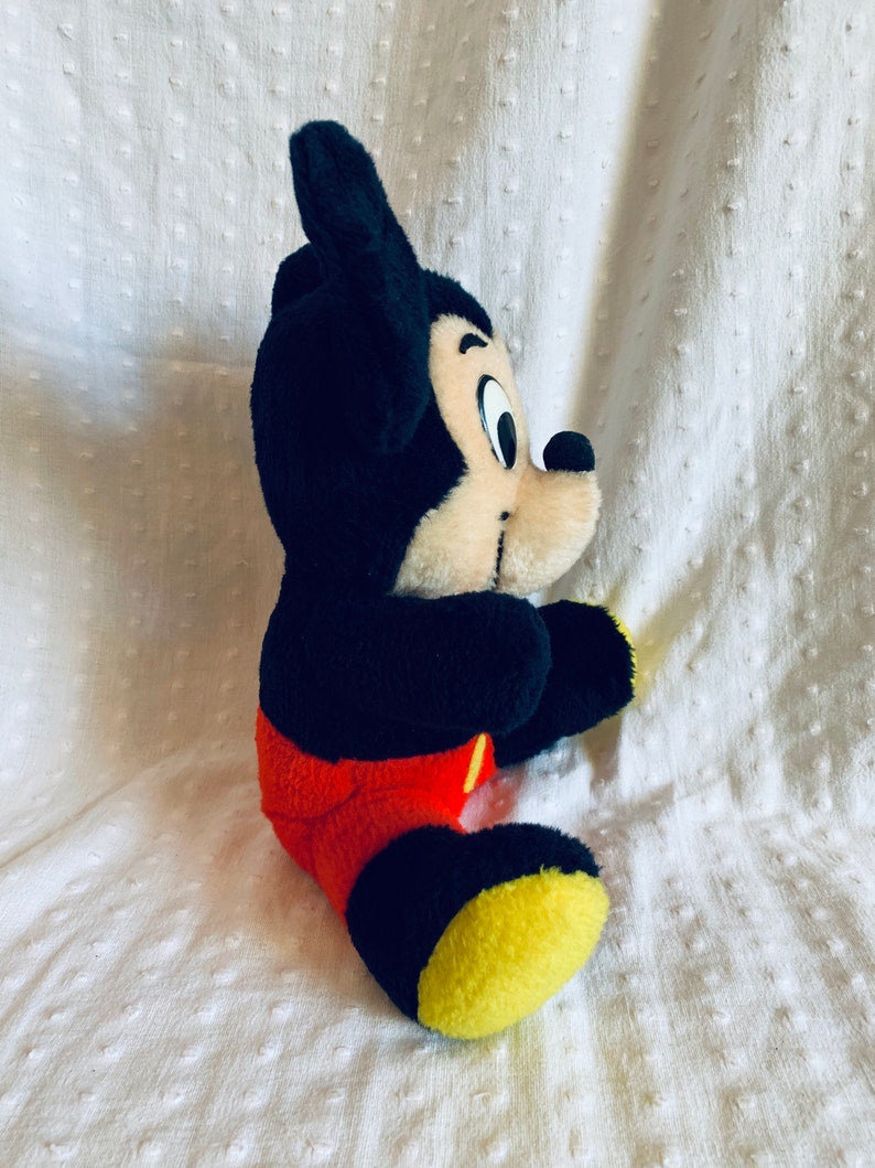 May include: A plush Mickey Mouse toy sitting on a white surface. The toy is black with red shorts and yellow shoes. Mickey Mouse has a black nose and large black ears.