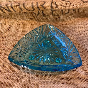 May include: A triangular, light blue glass dish with a decorative floral pattern. The dish has a starburst design in the center and three small, round feet. The edges are slightly curved, and the dish is set against a burlap background.