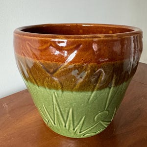 Könnte beinhalten: Ein brauner und grüner Keramiktopf mit einem strukturierten Design. Der Topf hat eine breite Öffnung und einen abgerundeten Boden.
